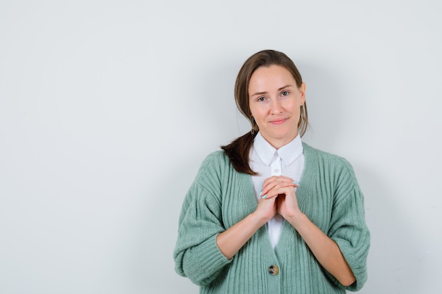 Portrait of young woman keeping clasped hands over chest in blouse, cardigan and looking cheery front view