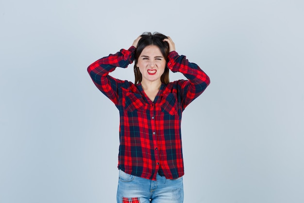 Portrait of young woman keeping hands on head in checked shirt, jeans and looking annoyed front view