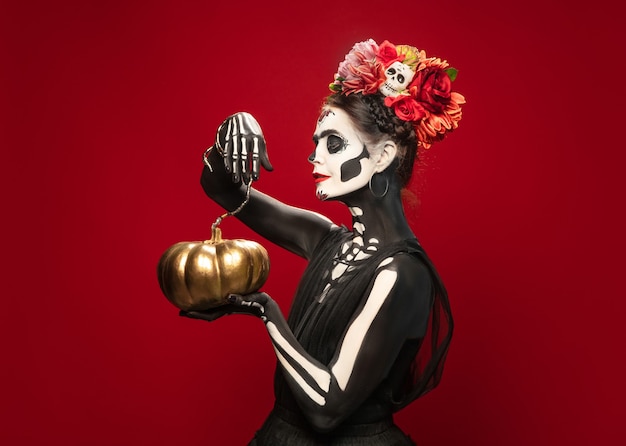 Portrait of young woman like santa muerte saint death or sugar skull with bright make-up. female isolated on red studio background with copyspace. celebrating halloween or day of the dead