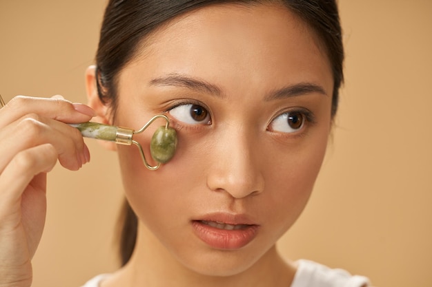 Portrait of young woman looking aside while using jade roller for massaging her face posing