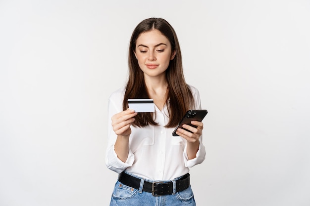 Portrait of young woman looking at smartphone screen, paying for purchase online with credit card, standing over white background, placing order.