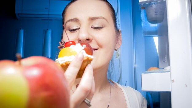 Ritratto di giovane donna che guarda la torta dolce in frigorifero durante la notte. concetto di dieta e alimentazione sana.