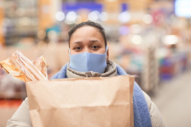 Портрет молодой женщины в маске, смотрящей в камеру, держащую хозяйственные сумки в супермаркете