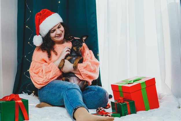 Portrait of young woman playing with funny dog miniature pinscher around Christmas ball and gift boxes