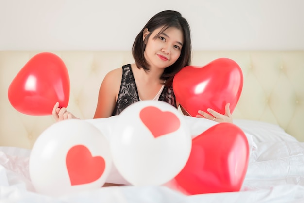 Portrait of young woman playing with white and red heart-shaped balloons on the bed