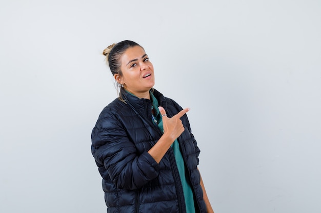 Portrait of young woman pointing at upper right corner in puffer jacket and looking confident front view