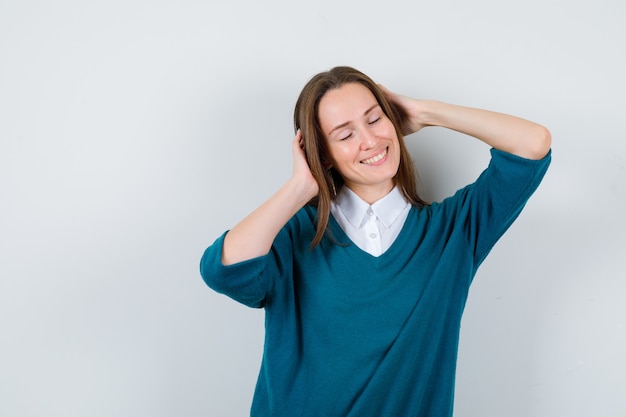 Portrait of young woman posing while keeping hands on head, shutting eyes in sweater over white shirt and looking merry front view