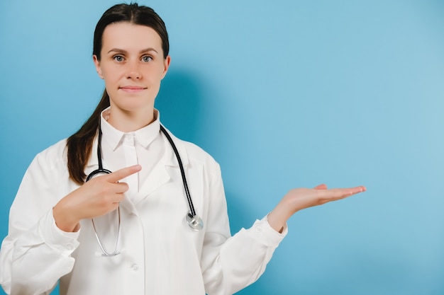 Portrait of young woman in professional medical white coat indicates with hands on copy space for advertisement. Doctor presenting and showing something isolated over blue studio background wall