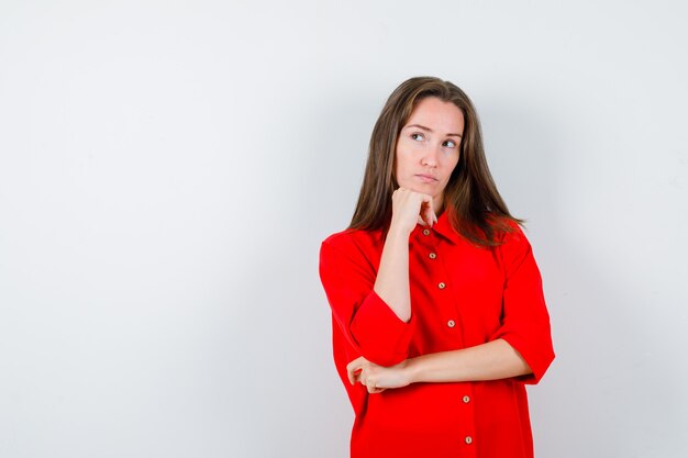 Portrait of young woman propping chin on hand in red blouse and looking thoughtful front view