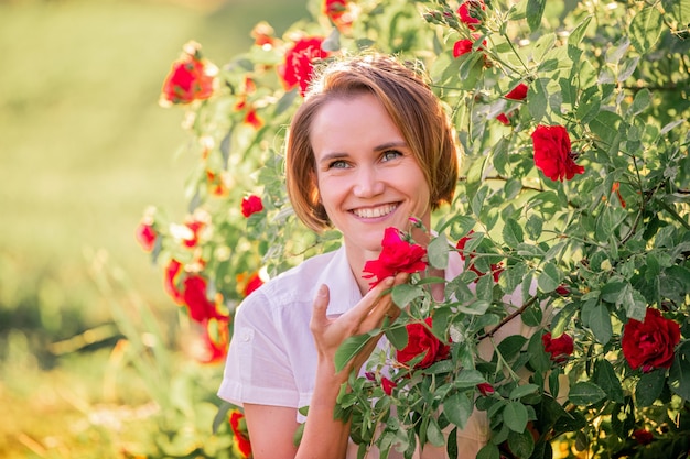 Portrait of a young woman among red roses in the back sunlight