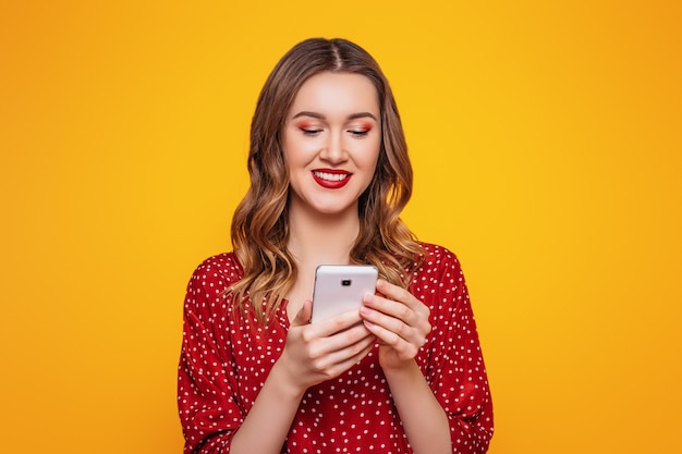 Portrait of a young woman in a red summer dress with red lips chatting by mobile phone isolated over orange wall wall. Online orders purchases shopping credit concept