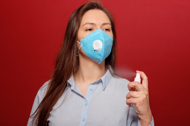 Portrait of young woman on red wall in protective medical mask sprays antiseptic on her face