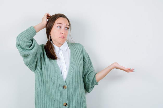 Portrait of young woman scratching head, showing helpless gesture in blouse, cardigan and looking clueless front view