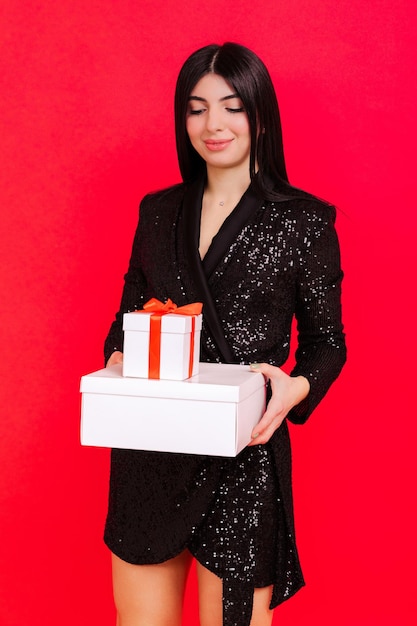 Portrait of a young woman in a shiny elegant dress holding boxes with gifts on a red background