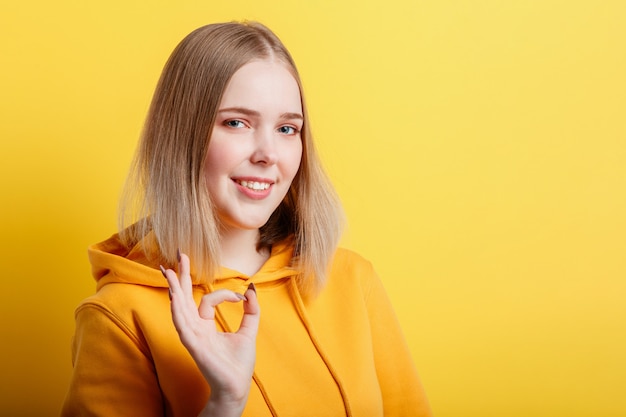 Portrait young woman shows ok hand gesture. Teenage blonde calm smiling girl shows positive ok gesture symbol like and approval isolated on color yellow background in Studio.