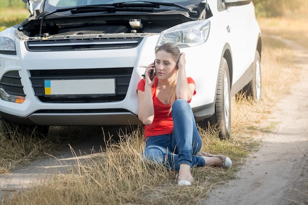 Portrait of young woman sitting next to broken car and calling for help