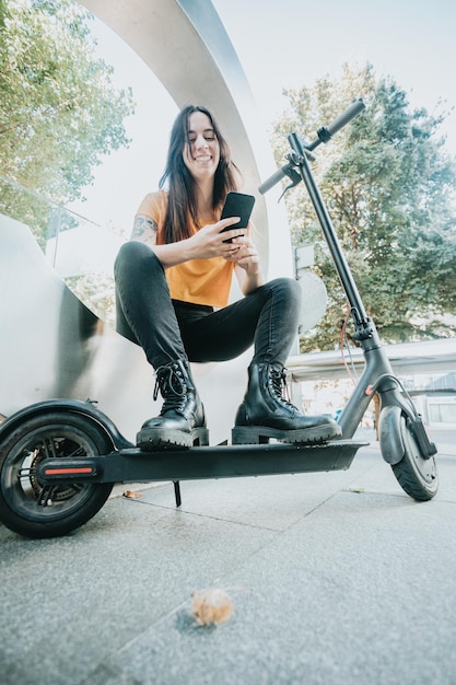 Portrait of young woman sitting on urban scenery holding a\
mobile phone and leaning on an electric scooter with a smile\
resting and watching social media with casual clothes city\
lifestyle