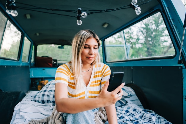 Portrait of young woman sitting in a van and using mobile phone, outdoors in nature. Enjoying summer, travel concept