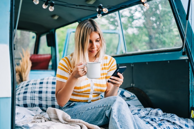 Portrait of young woman sitting in a van and using mobile phone, outdoors in nature. Enjoying summer, travel concept