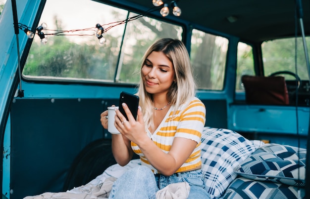 Portrait of young woman sitting in a van and using mobile phone, outdoors in nature. Enjoying summer, travel concept