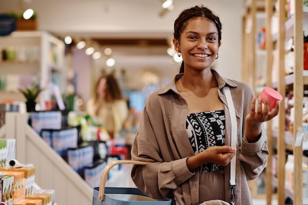 Portrait of young woman smiling at camera while choosing cream for herself in the store