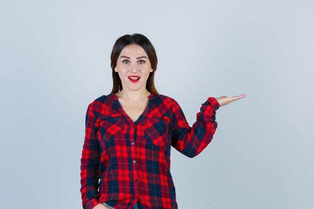 Portrait of young woman spreading palm aside in checked shirt and looking amazed front view