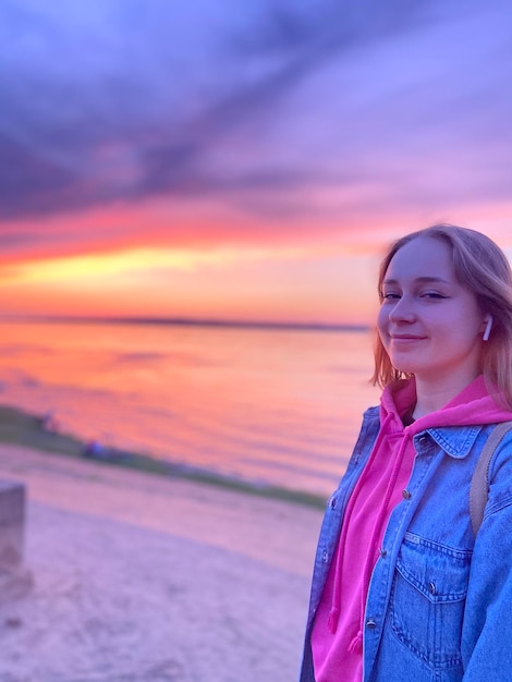 Portrait of young woman standing at beach against sky during sunset