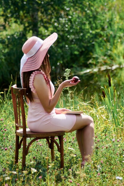 Portrait of a young woman in a straw hat on a background of greenery. She is sitting with her back to the camera and holding an apple in her hand.