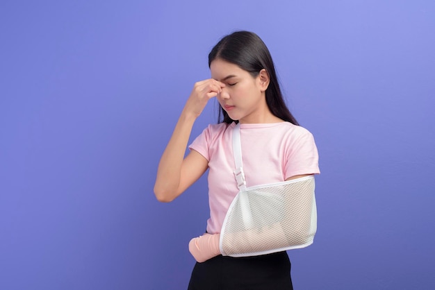 A portrait of young woman stressed and frustrated with an injured arm in a sling over blue background in studio, insurance and healthcare concept