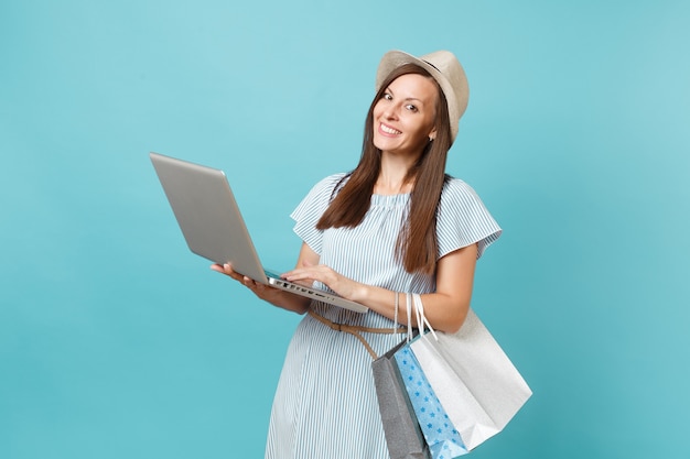 Portrait of young woman in summer dress, straw hat holding packages bags with purchases after online shopping, using laptop pc computer isolated on blue pastel background. Copy space for advertisement