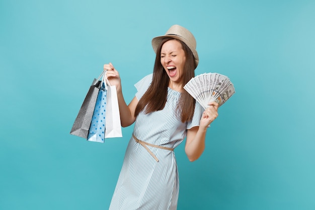 Portrait young woman in summer dress, straw hat holding packages bags with purchases after shopping, lots of dollars banknotes, cash money, isolated on blue background. Copy space for advertisement.