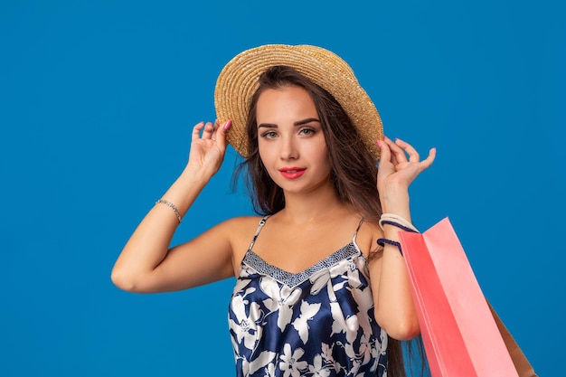 Portrait of a young woman in summer hat holding shopping bags and looking at the camera isolated over blue background