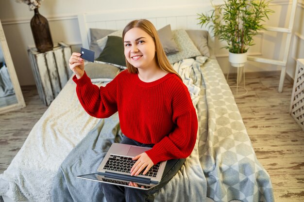 Ritratto di una giovane donna in un maglione, utilizzando un laptop e una carta di credito, seduta a casa sul letto, shopping online, internet banking. acquisti a domicilio