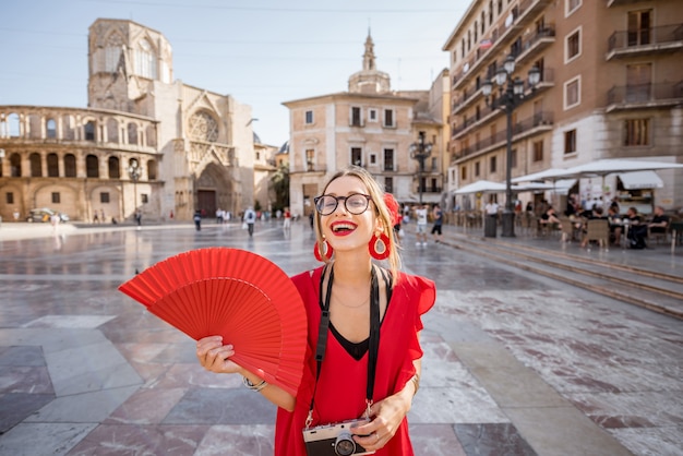 Portrait of a young woman tourist in red dress with hand fan on the central square of the old town in Valencia city, Spain