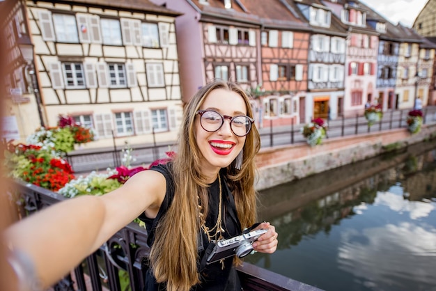 フランス北東部のアルザス地方のコルマール村の古い橋の上に立っている写真カメラを持つ若い女性観光客の肖像画