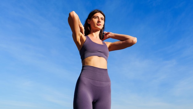 青い空を背景に夏の外でトレーニングする前に、ヘッドフォンでトラック スーツを着た若い女性の肖像画