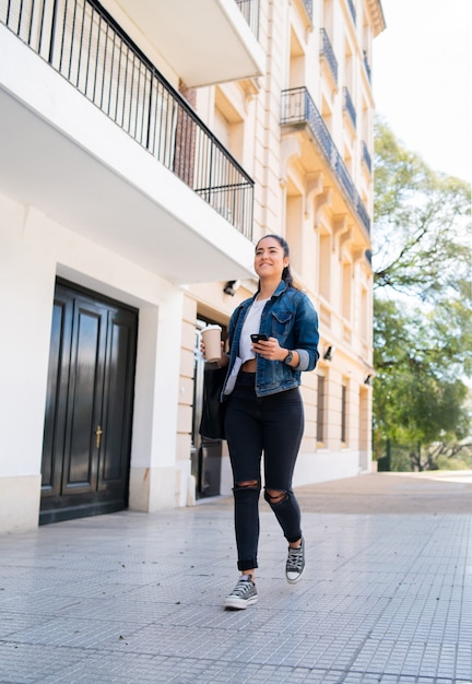 Ritratto di giovane donna che utilizza il suo telefono cellulare e che tiene una tazza di caffè mentre cammina all'aperto sulla strada. concetto urbano.