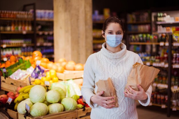 Portrait of young woman in warm sweatre wearing protective mask holding two paper bags with food Shelves with various products on the background Supporting local business concept