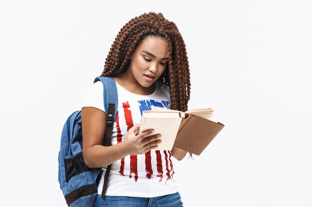 Portrait of young woman wearing backpack reading book while studying in college standing isolated against white wall