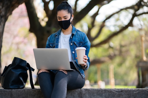 Portrait of young woman wearing face mask and using her laptop while sitting outdoors. Urban concept.