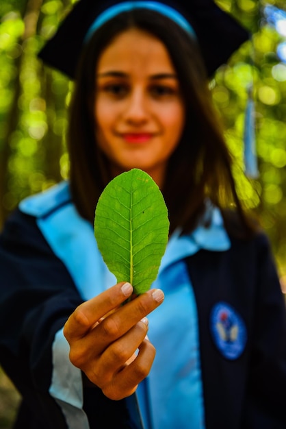 Foto ritratto di una giovane donna che indossa un abito da laurea mentre tiene una foglia all'aperto