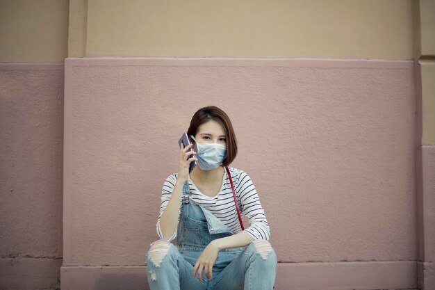 Portrait of young woman wearing mask talking over smart phone while sitting against wall