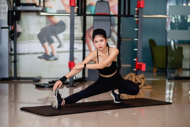 Ritratto di giovane donna che indossa abbigliamento sportivo e smartwatch seduto sul pavimento e allungando i muscoli delle gambe e delle braccia prima dell'allenamento in palestra,