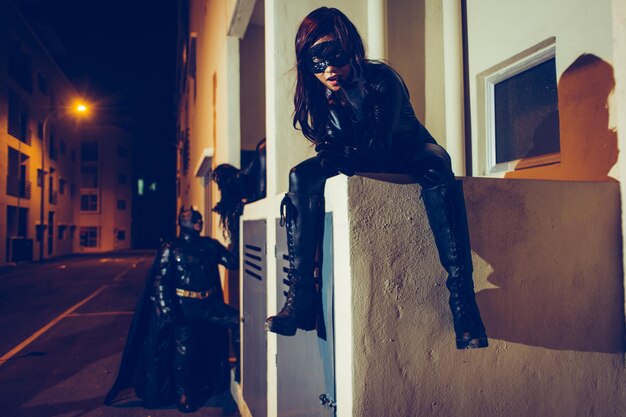 Photo portrait of young woman wearing superhero costume sitting against building at night