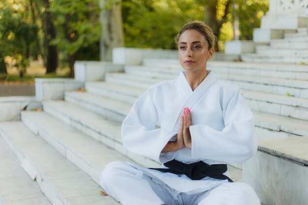 Portrait of young woman in white kimono with black belt. Sport woman sitting on stairs and meditating outdoors. Martial arts