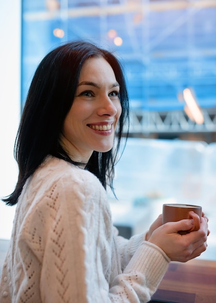 카페에 있는 동안 손에 커피 한 잔을 들고 하얀 스웨터를 입은 젊은 여성의 초상화