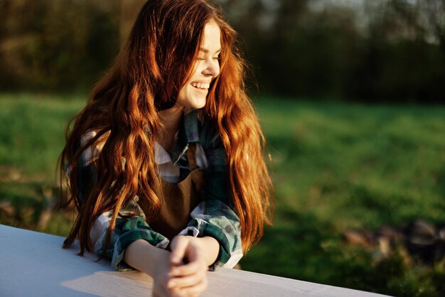 Photo portrait of a young woman with beautiful long red hair with a beautiful smile with teeth against a green park in the summer sunset sunlight