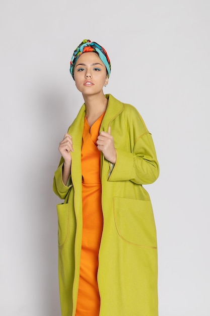 Portrait of a young woman with bright makeup and a fashionable headscarf light background beauty fashion makeup concept girl in a bright green coat bright orange dress 169 panoramic format