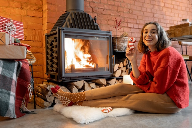 Portrait of a young woman with a Christmas candy by the fireplace. House coziness and warmth during winter holidays