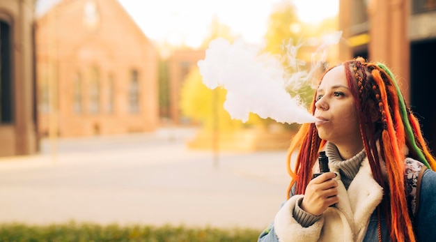 Portrait of young woman with dreadlocks vaping standing on street Female with colourful hairstyle smoking ecigarette letting off steam